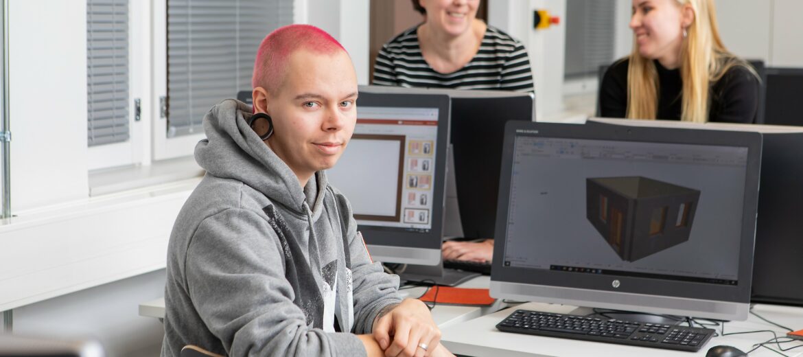 Punahiuksinen opiskelija istuu hymyillen tietokoneen äärellä.