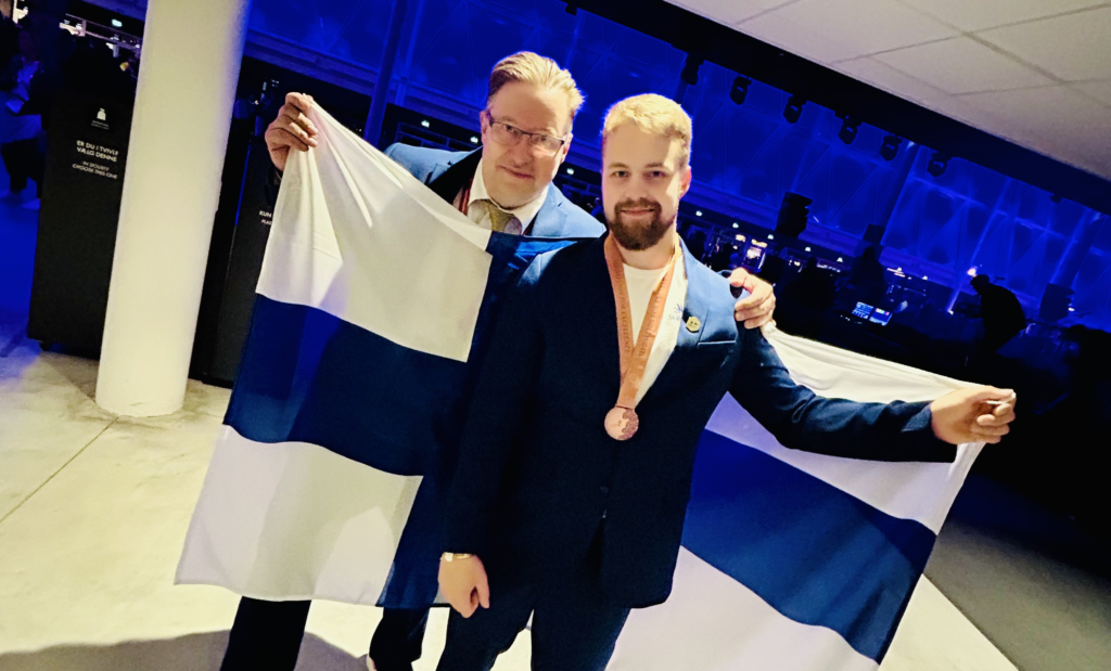 Ekspertti Tommi Mäntylä sekä kilpailija Viljami Turve katsovat kameraan pitäen Suomen lippua.
