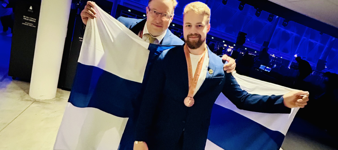 Ekspertti Tommi Mäntylä sekä kilpailija Viljami Turve katsovat kameraan pitäen Suomen lippua.