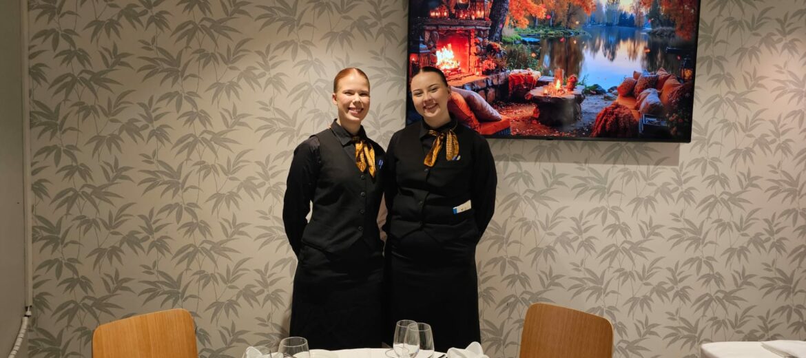 Tarjoilijaopiskelijat Oona Mäntyniemi ja Aino Ahvonen katsovat kameraan hymyillen.