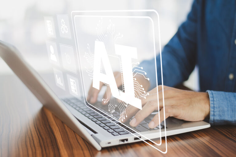 henkilö työskentelee kannettavalla tietokoneella. Kuvassa näkyy kannettava tietokone ja kädet sekä teksti AI.